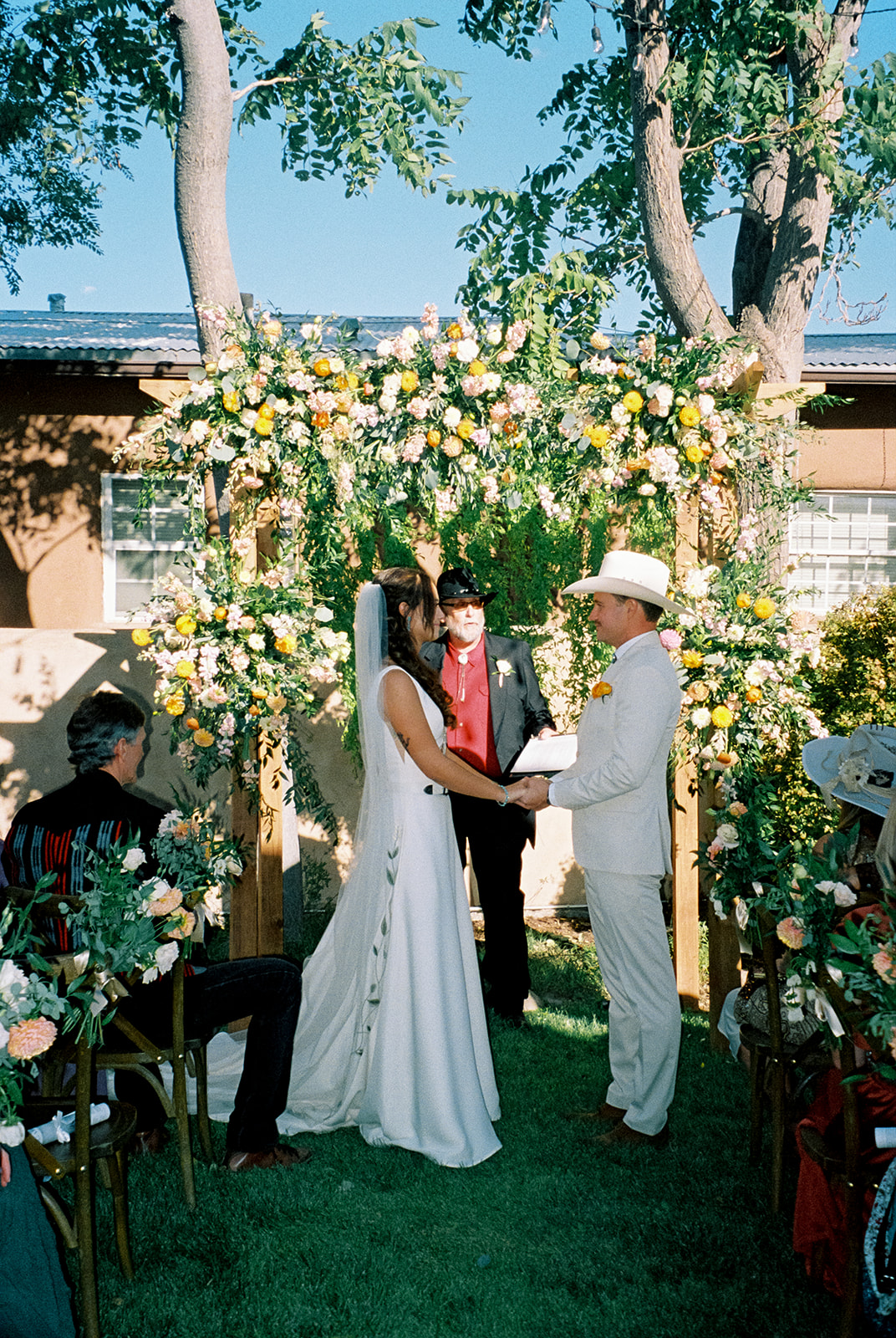 Bride and groom hold hands during western wedding ceremony, surrounded by a wildflower floral arch.