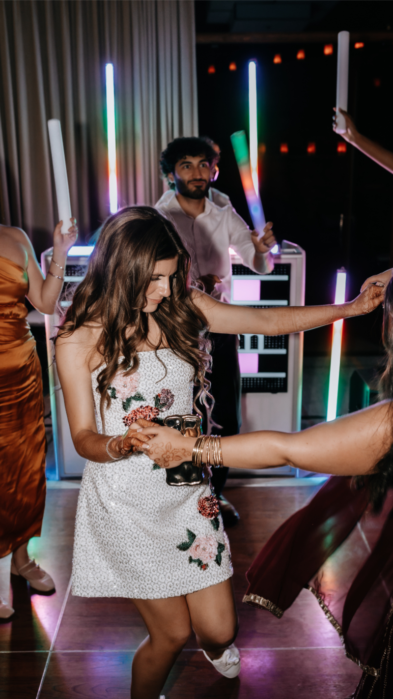 Bride dancing in a floral beaded reception dress, holding glow sticks with guests on a colorful lit dance floor.