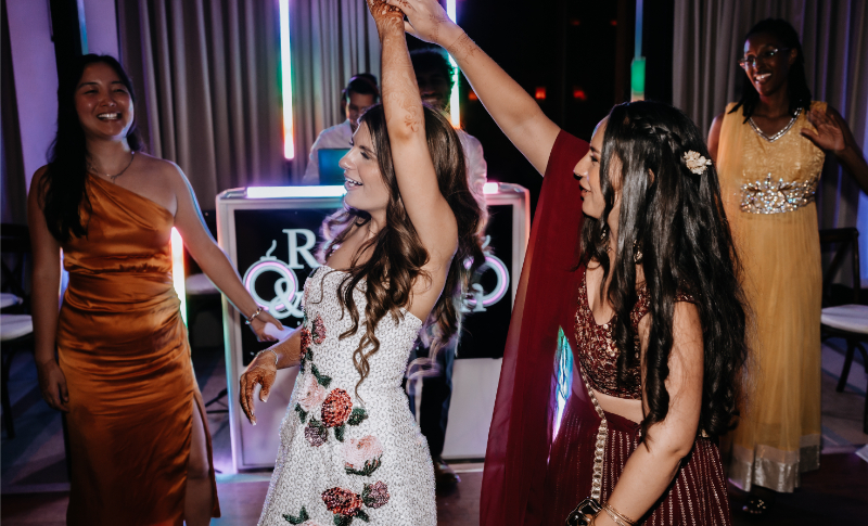Bride in a white reception dress raises her arms to dance with a guest in a red lehenga under a string-lit sign.