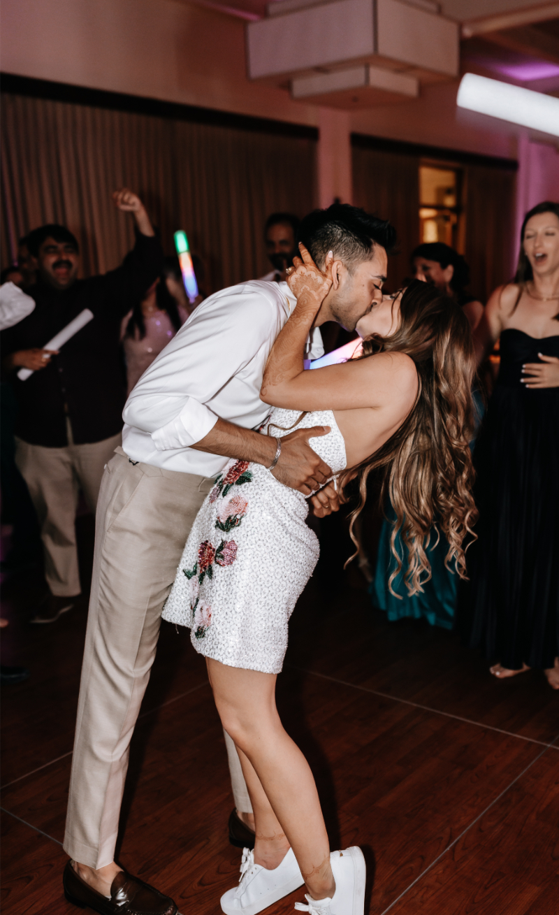 Bride and groom kiss on the dance floor as guests cheer, her short reception dress and his rolled sleeves suited to a night of dancing.