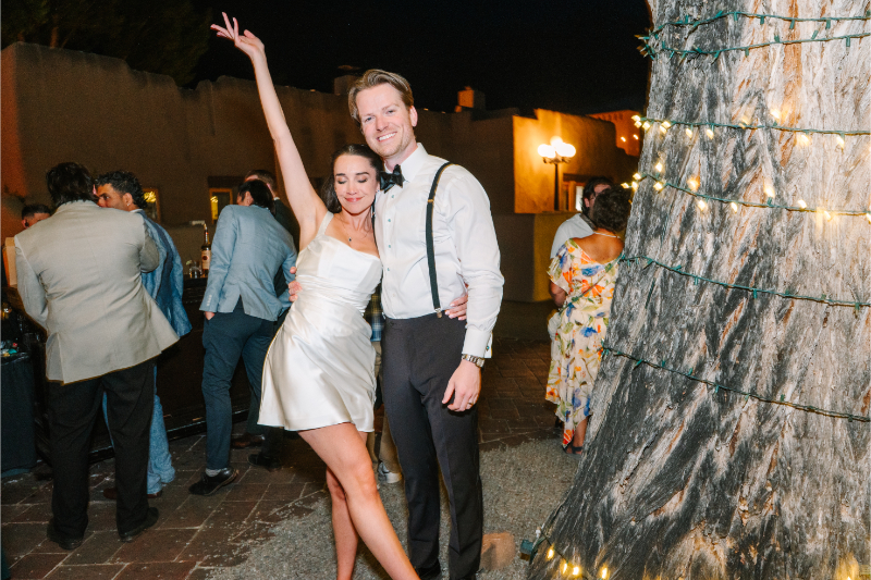Bride in a satin reception dress kicks up her leg for a playful photo with the groom under string lights.