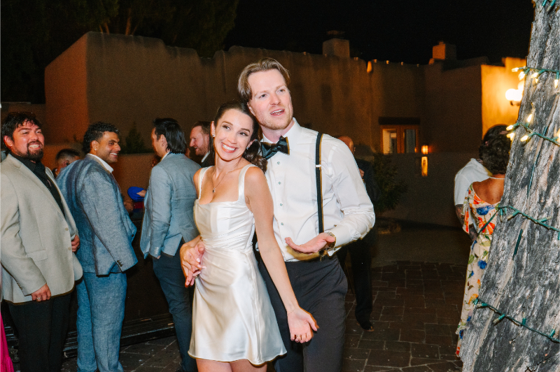 Bride in a satin reception dress kicks up her leg for a playful photo with the groom under string lights.