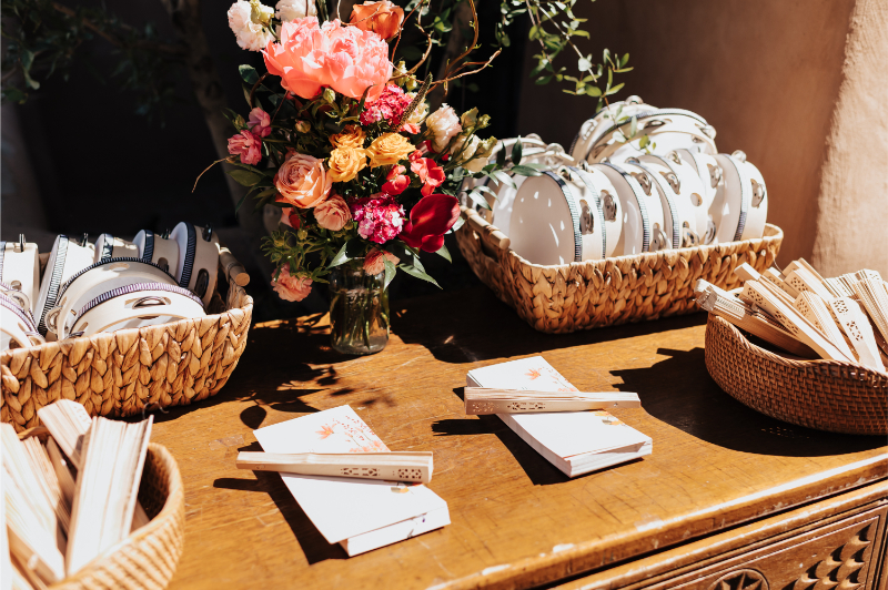 Wedding welcome table with baskets of striped napkins, patterned plates, and paper fans surrounding a coral and orange floral arrangement in a glass vase.