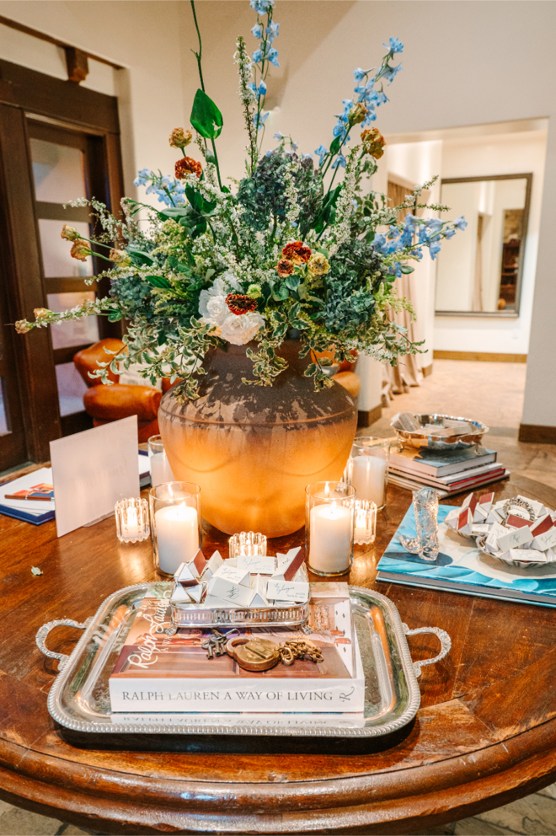 Wedding welcome table styled with a wild floral arrangement in a ceramic vase, a Ralph Lauren coffee table book, candles, and triangular match book favor boxes on a silver tray.