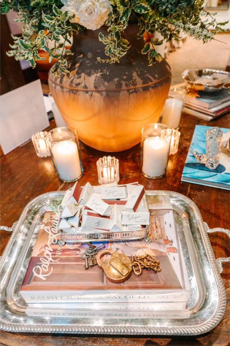 Silver tray holding a Ralph Lauren book, antique keys and lock, and triangular wedding favor boxes, set beside glowing candles.