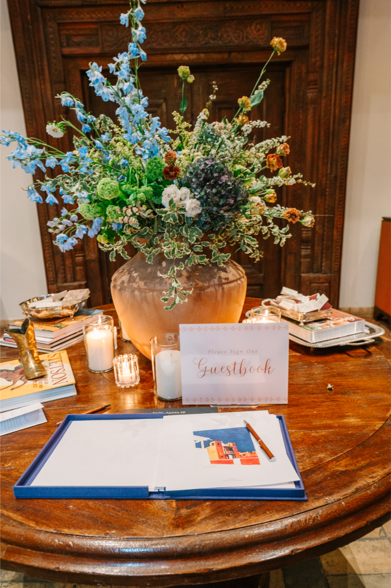 Wedding welcome table with a wildflower arrangement, a "Please Sign Our Guestbook" sign, and an open guestbook illustrated with adobe architecture.