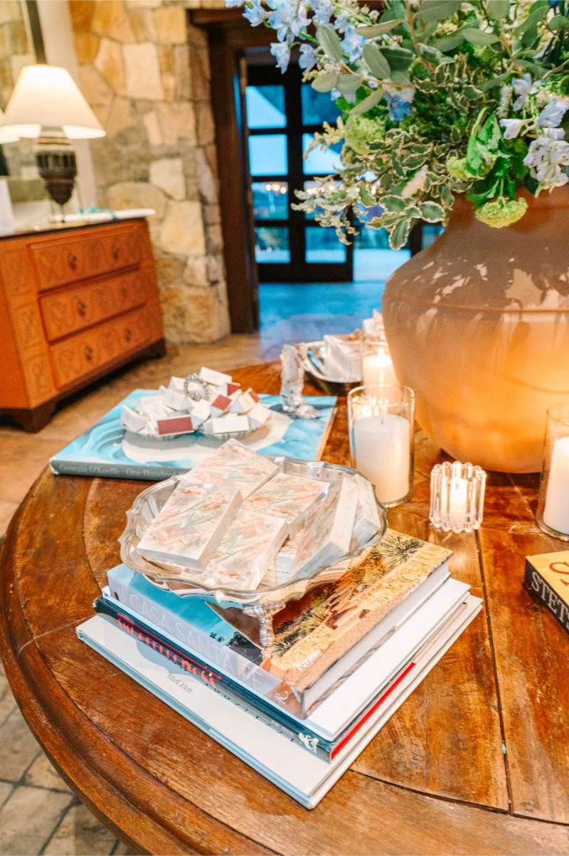 Wedding welcome table styled with stacked coffee table books, triangular favor boxes, candles, and a lush floral centerpiece.