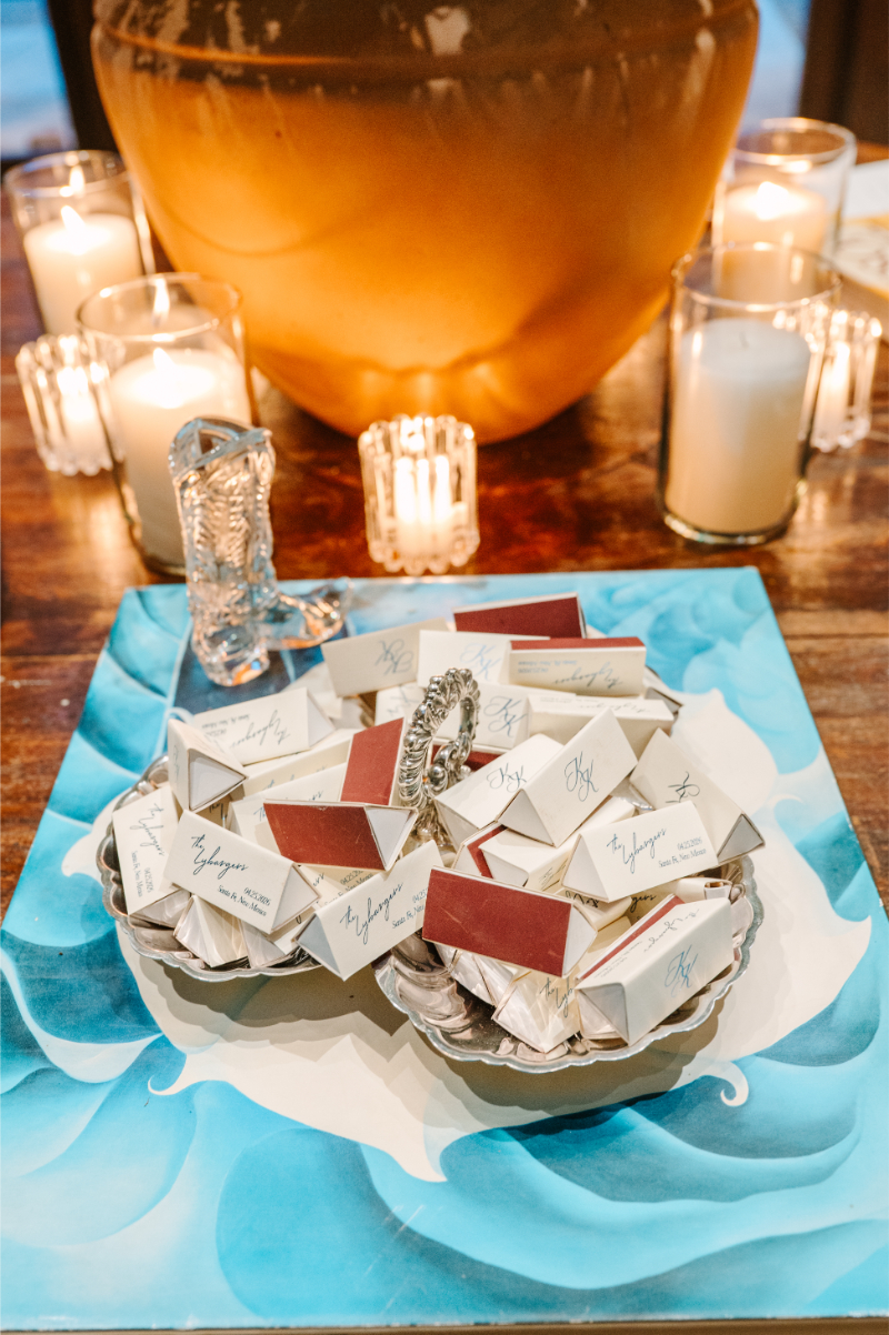 Close-up of personalized triangular wedding favor boxes arranged on silver trays atop a coffee table book, with candles and a silver boot accent.