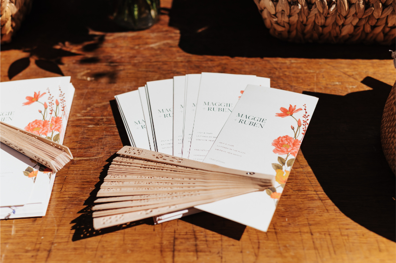 Close-up of ivory wedding fans printed with "Maggie + Ruben" and a coral floral design, fanned open on a wooden welcome table.