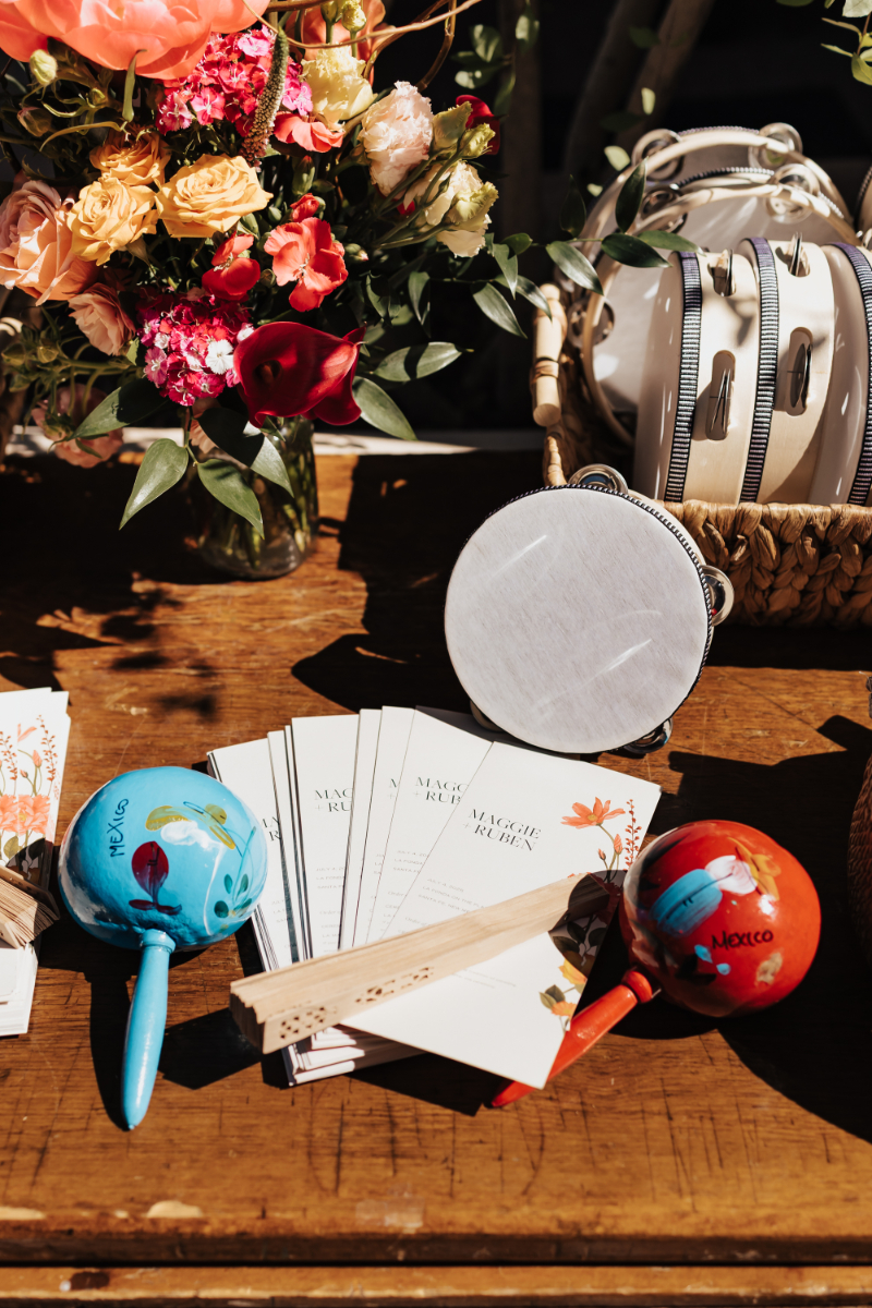 Blue and red maracas painted "Mexico" next to tambourines, paper fans, and a floral arrangement on a wedding welcome table.