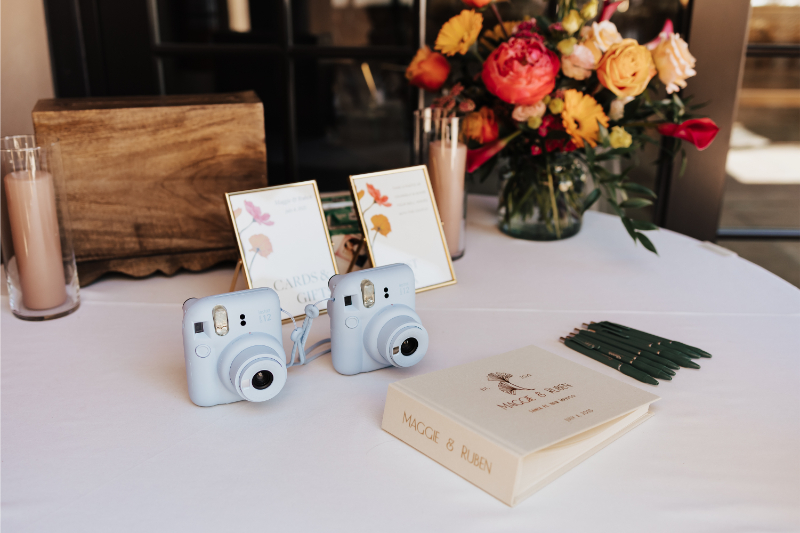 Wedding welcome table set with two white instant cameras, a personalized ivory guest book, green pens, and framed signage.