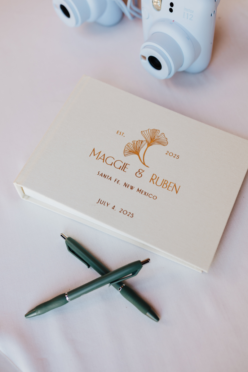 Close-up of a gold foil wedding guest book reading "Maggie & Ruben, Santa Fe, New Mexico, July 4, 2025" with crossed green pens and instant cameras nearby.