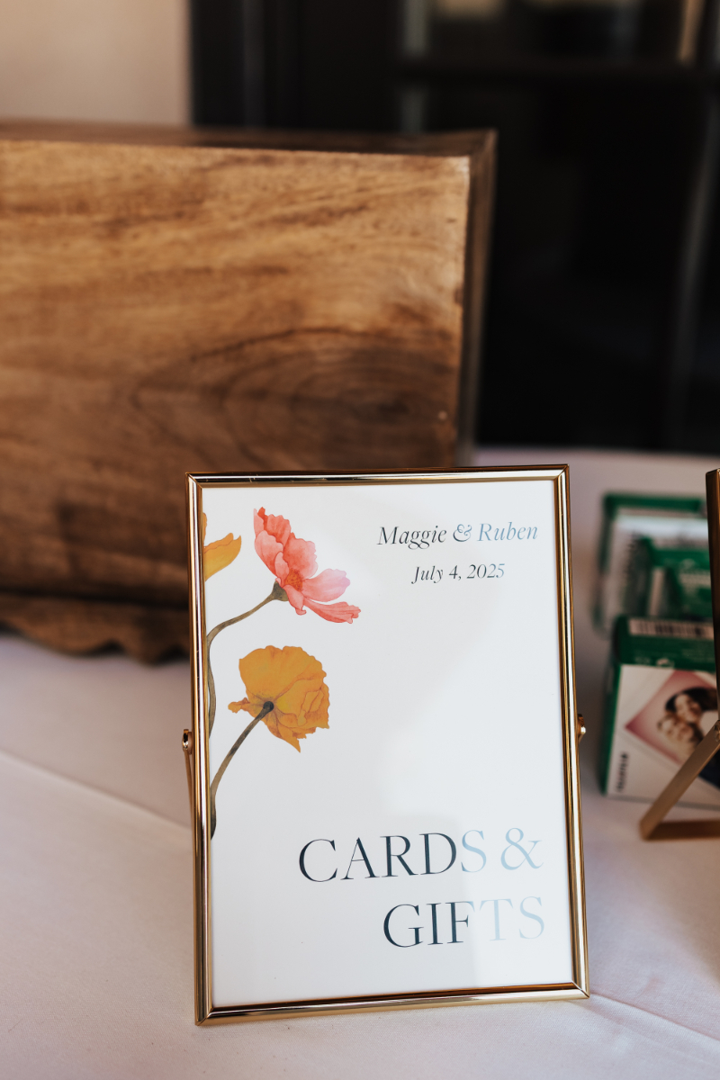 Gold-framed "Cards & Gifts" sign with a watercolor floral design on a wedding welcome table.